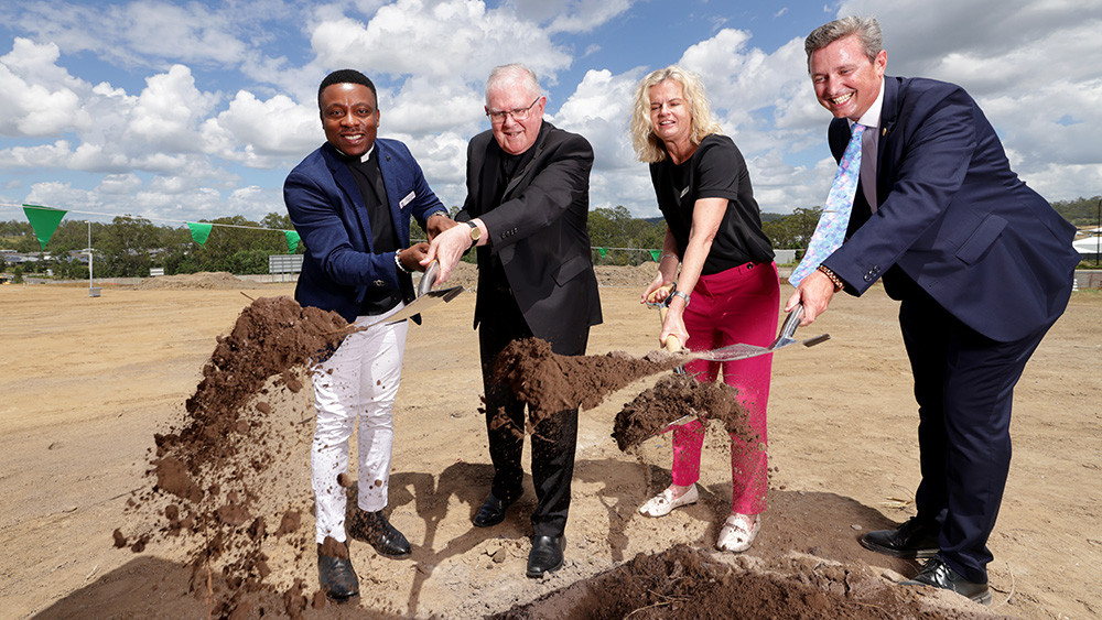 Four adults using shovels to toss soil during a ceremonial ground‑breaking event at a construction site
