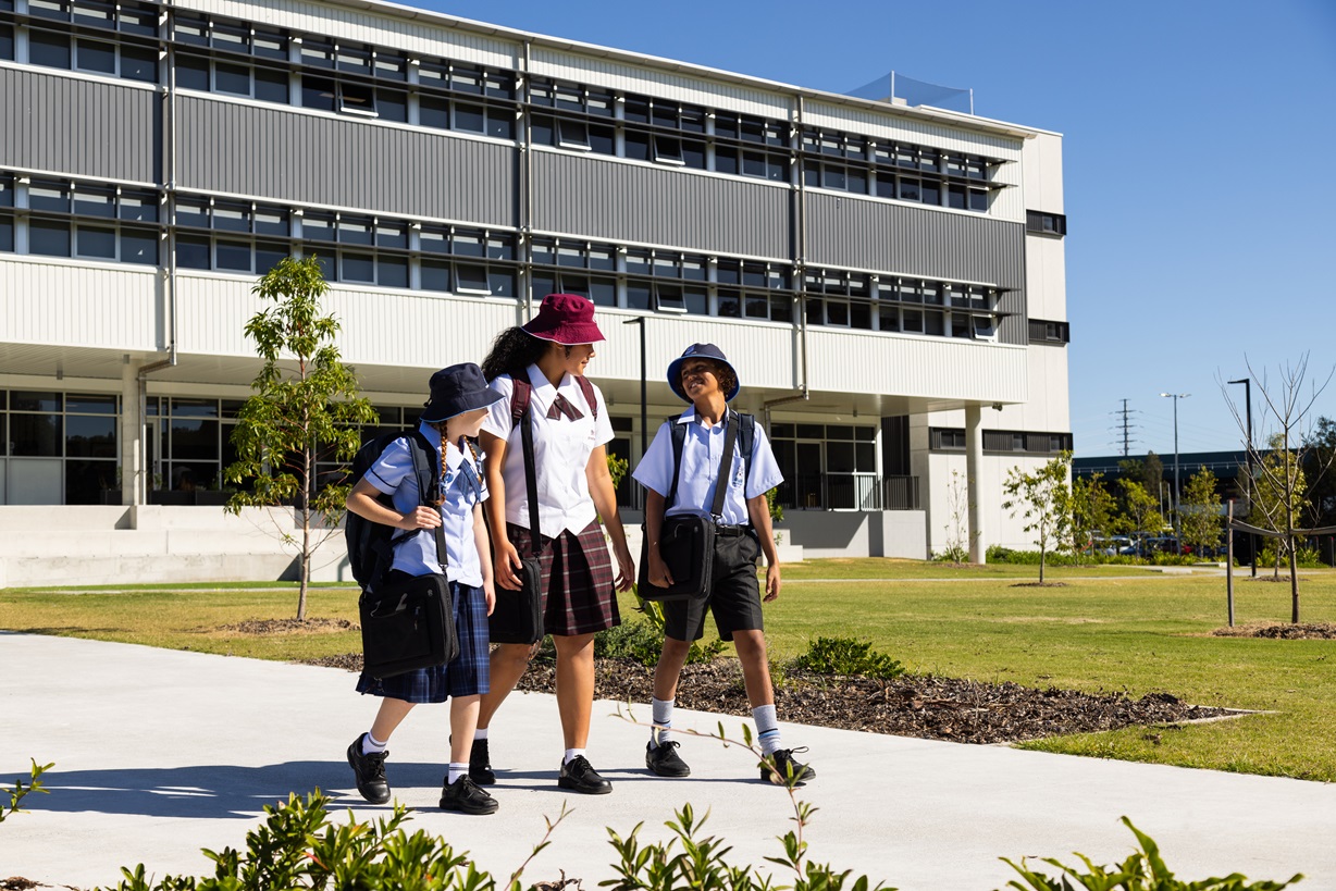 Three students in school uniforms walking along a path in front of a modern multi-storey school building