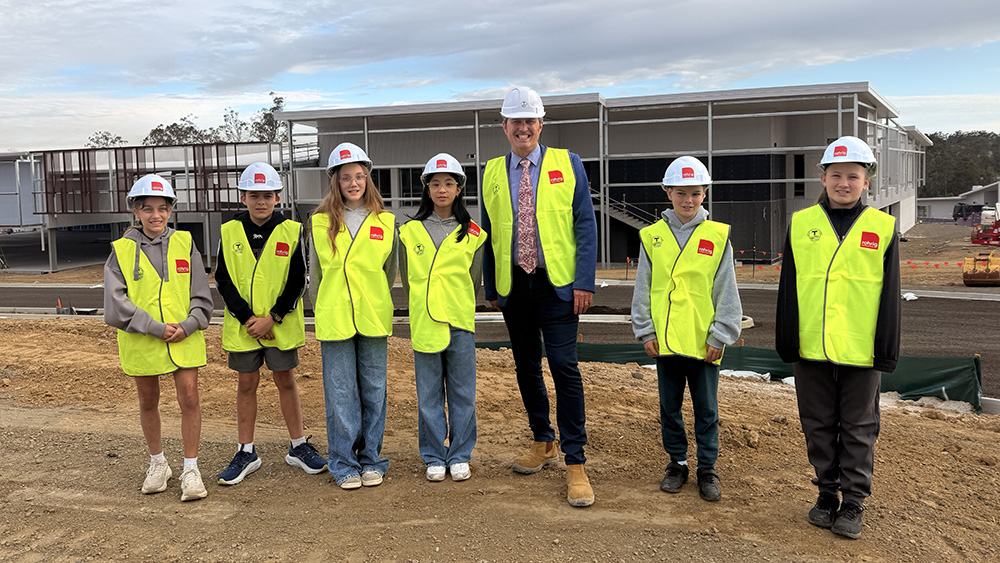 Students and an adult wearing high-visibility vests and hard hats standing in front of a school building under construction